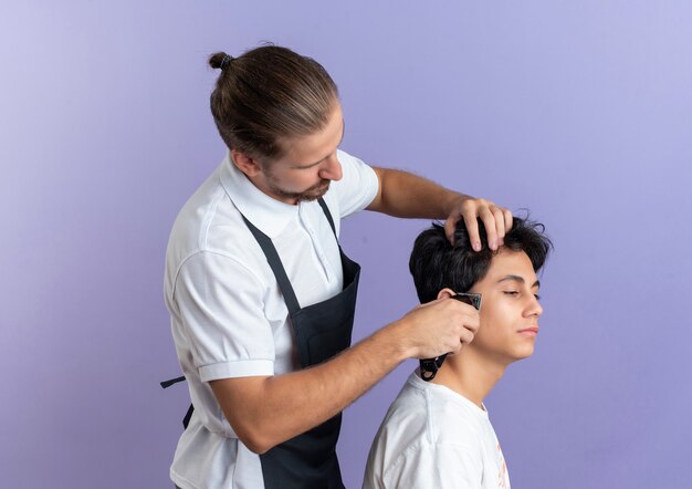 Young handsome barber wearing uniform standing in profile view doing haircut for young client isolated on purple background with copy space