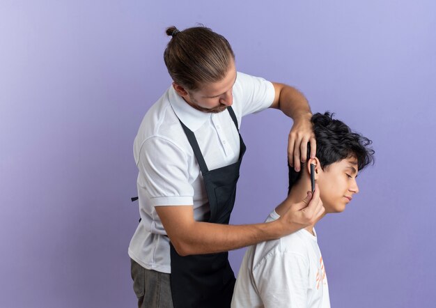 Young handsome barber wearing uniform standing in profile view doing haircut for young client isolated on purple background with copy space