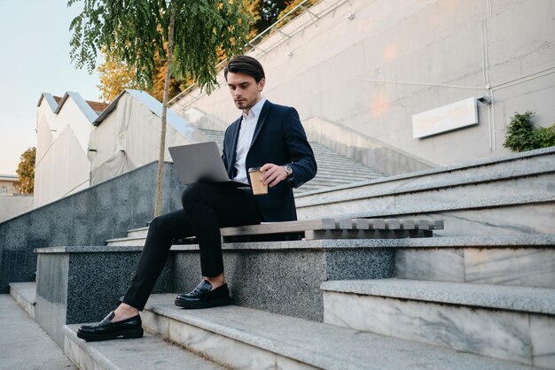 Young handsome bearded brunette man in white shirt and classic suit sitting on bench with cup of coffee to go in hand thoughtfully working on laptop alone