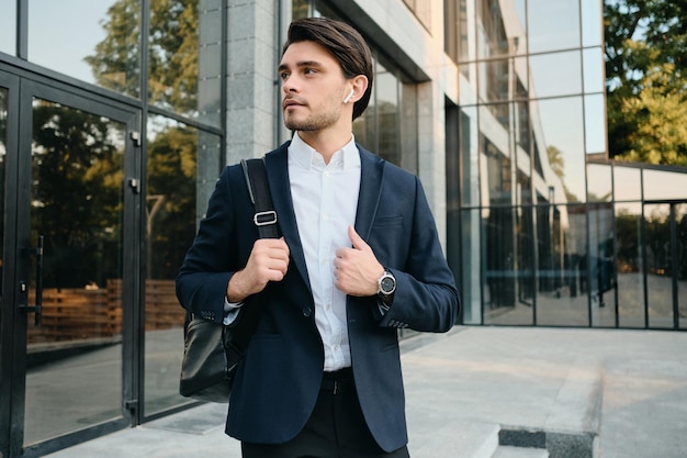 Young handsome bearded brunette man in white shirt and classic suit with wireless earphones and backpack on shoulder dreamily looking aside with glass building on background