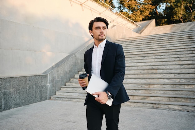 Young handsome bearded brunette man in white shirt and classic suit with wireless earphones holding cup of coffee to go and laptop in hands thoughtfully looking aside in city park