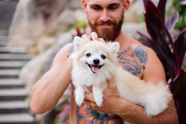 Young handsome bearded brutal tattooed happy man holds pomeranian spitz playing with lovely pet