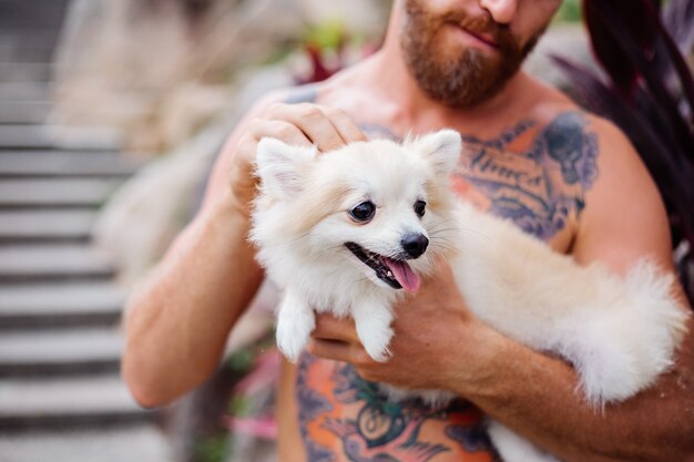 젊은 잘 생긴 수염 난 잔인한 문신을 한 행복한 사람은 사랑스러운 애완 동물을 가지고 노는 포메라니안 스피츠를 보유하고 있습니다.