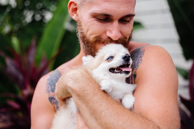 Молодой красивый бородатый брутальный татуированный счастливый мужчина держит померанского шпица, играя с прекрасным домашним животным