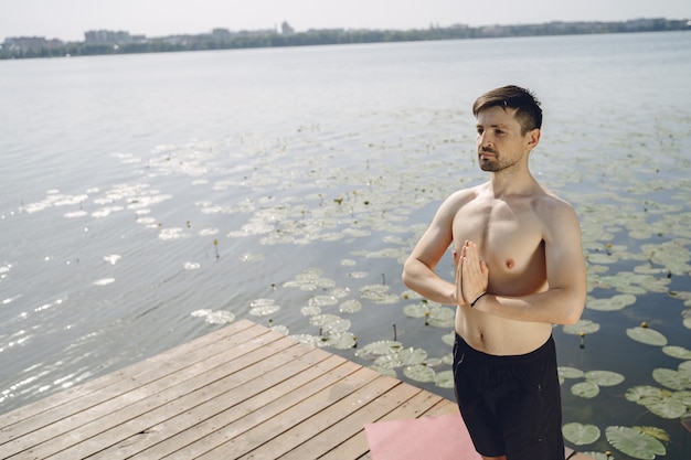 Young handsome bearded man sitting on wooden pier in summer day. Meditating and relaxing.