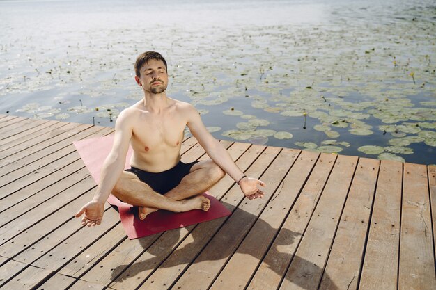 夏の日に木製の桟橋に座っている若いハンサムなひげを生やした男。瞑想とリラックス。