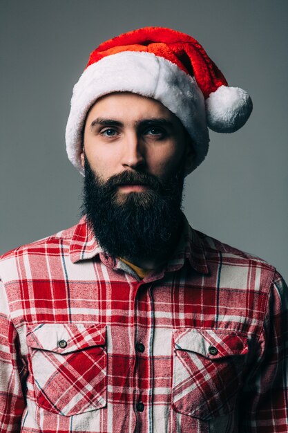 Молодой красивый бородатый мужчина с красной рождественской шляпой на серой стене