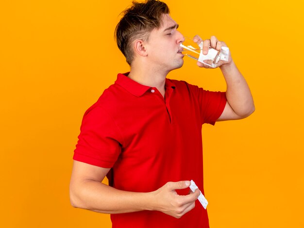 Молодой красивый блондин больной мужчина держит салфетку, принимая пилюлю питьевой воды из стекла, изолированного на оранжевой стене