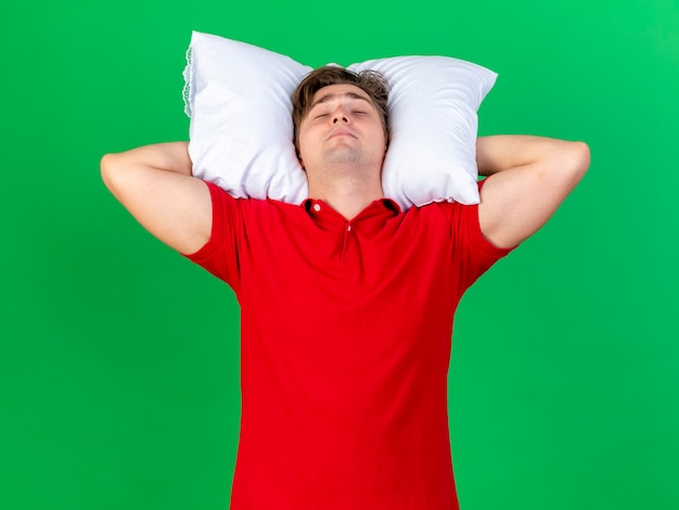 Free photo young handsome blonde ill man holding pillow under head pretend sleeping isolated on green background