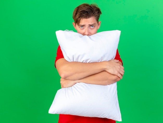 Young handsome blonde ill man holding pillow looking at camera from behind it isolated on green background with copy space