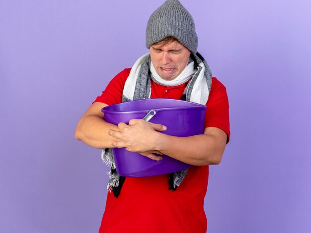 冬の帽子とスカーフを身に着けている若いハンサムな金髪の病気の男は、コピースペースで紫色の背景に分離された目を閉じて吐き気を持っているプラスチック製のバケツを保持しています