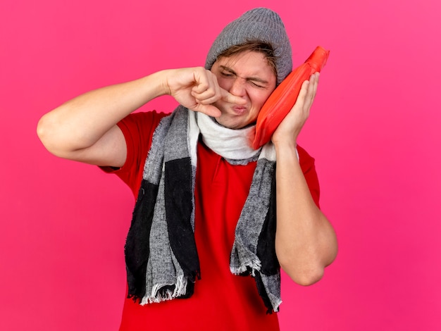 Young handsome blonde ill man wearing winter hat and scarf touching face with hot water bottle wiping nose with finger with closed eyes isolated on crimson background with copy space