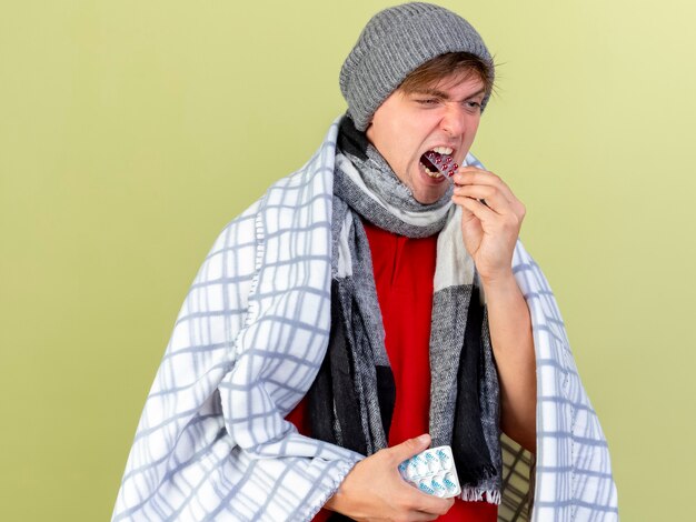 Young handsome blonde ill man wearing winter hat and scarf wrapped in plaid holding packs of medical pills and putting it in mouth looking straight isolated on olive green wall