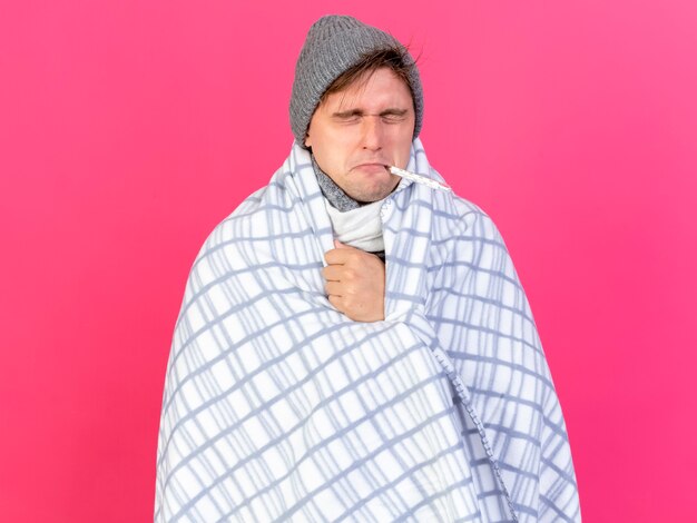 Young handsome blonde ill man wearing winter hat and scarf wrapped in plaid holding thermometer in mouth with closed eyes isolated on crimson background with copy space