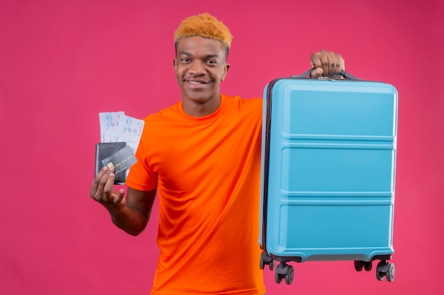 Free photo young handsome boy wearing orange t-shirt holding travel suitcase and air tickets smiling confident positive and happy standing over pink wall