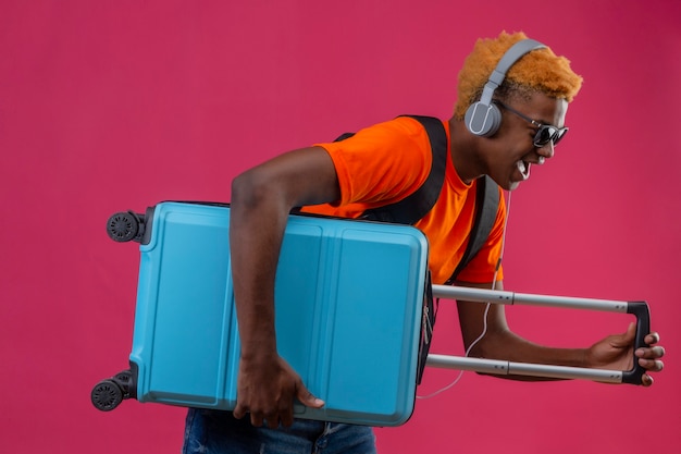 Молодой красивый мальчик в оранжевой футболке с наушниками на голове держит дорожный чемодан
