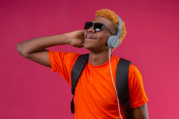 Young handsome boy with backpack and headphones enjoying favorite music happy and positive smiling standing over pink wall