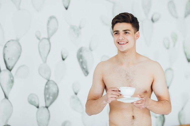 Giovane bel ragazzo con il torso nudo che beve caffè nella sua stanza al mattino