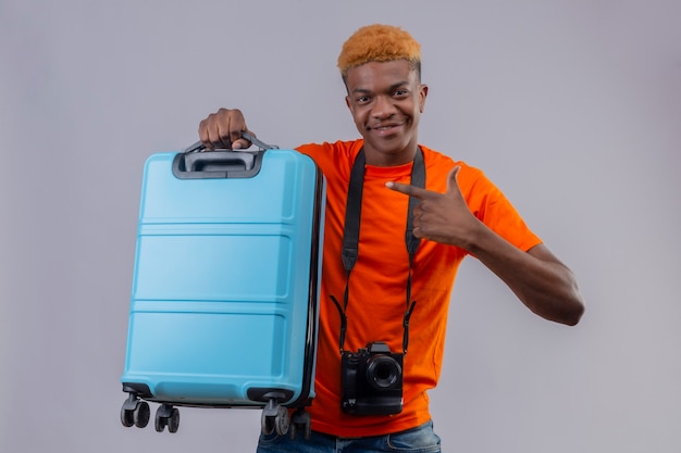 Young handsome boy with camera wearing orange t-shirt holding travel suitcase pointing with finger to it looking confident