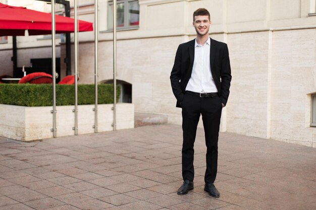 Young handsome businessman in black suit and white shirt with wireless earphones happily looking in camera while spending time on street