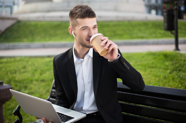クラシックな黒のジャケットと白いシャツを着た若いハンサムなビジネスマンがベンチに座ってラップトップを膝の上に置き、夢のように通りを脇に見ながらコーヒーを飲みます