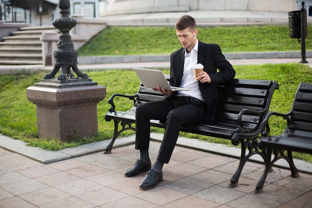 Giovane bell'uomo d'affari in classico abito nero e camicia bianca con auricolari wireless e tazza di caffè per andare in mano pensieroso lavorando sul laptop mentre si è seduti su una panchina all'aperto