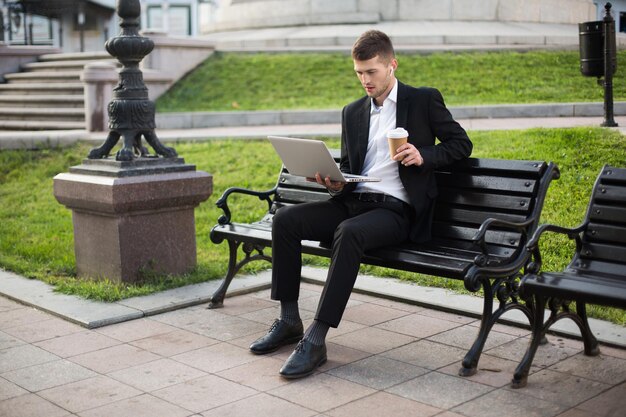 Young handsome businessman in classic black suit and white shirt with wireless earphones and cup of coffee to go in hand thoughtfully working on laptop while sitting on bench outdoor