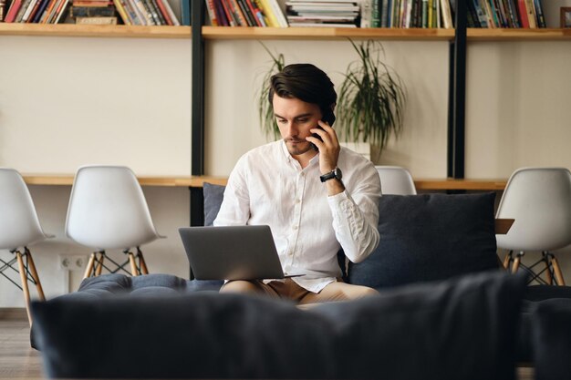 Young handsome businessman sitting on sofa thoughtfully talking on cellphone and working on laptop in modern coworking space