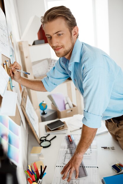 Young handsome businessman standing at table and writing on the paper pinned to corkboard.