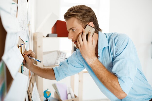 Молодой красивый бизнесмен, писать на бумаге, закрепленный на пробковой доске, разговаривает по телефону.