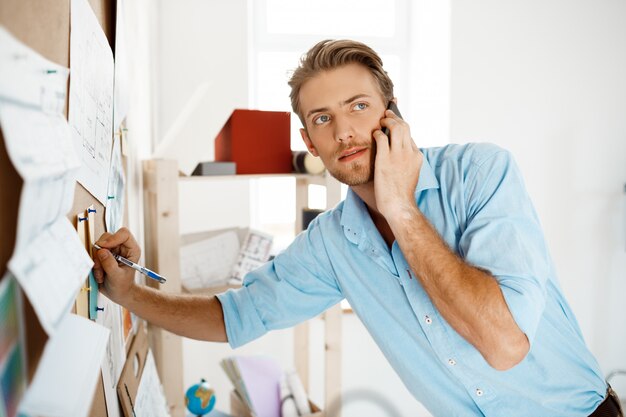 Young handsome businessman writing at the paper pinned to corkboard, talking on  phone.