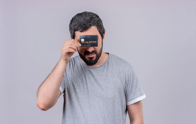 Молодой красивый кавказский мужчина держит и прячется за кредитной картой, изолированной на белом фоне с копией пространства