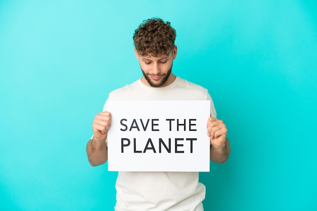 Молодой красивый кавказский мужчина, изолированные на синем фоне, держит плакат с текстом «спасите планету»