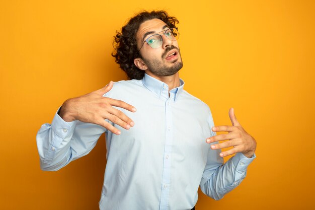 Young handsome caucasian man wearing glasses looking at camera keeping hands in air explaining something isolated on orange background