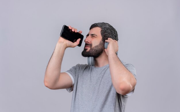 Young handsome caucasian man wearing headphones pretend singing and using his mobile phone as microphone with closed eyes and hand on headphone isolated on white background with copy space