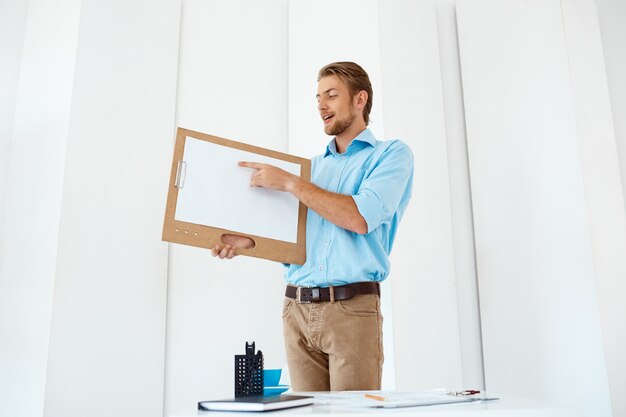 Молодой красивый жизнерадостный бизнесмен стоя на таблице держа деревянную доску сзажимом для бумаги с белым листом указывая на его. Светлый современный офисный интерьер