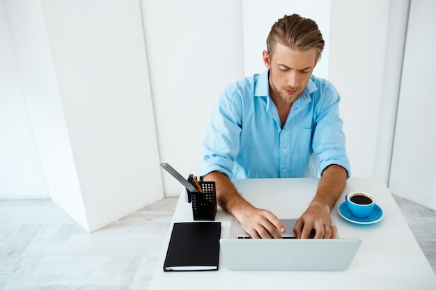 Free photo young handsome cheerful confident pensive businessman sitting at table working on laptop with cup of coffee aside. white modern office interior