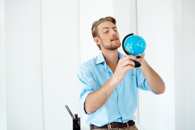 Young handsome cheerful confident pensive businessman standing at table holding small globe. White modern office interior