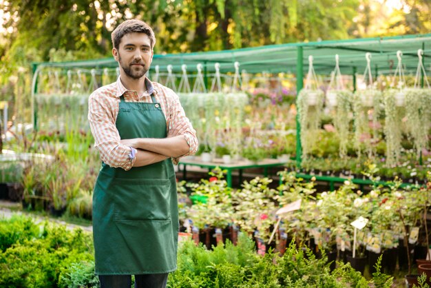 Young handsome cheerful gardener smiling, posing with crossed arms among flowers