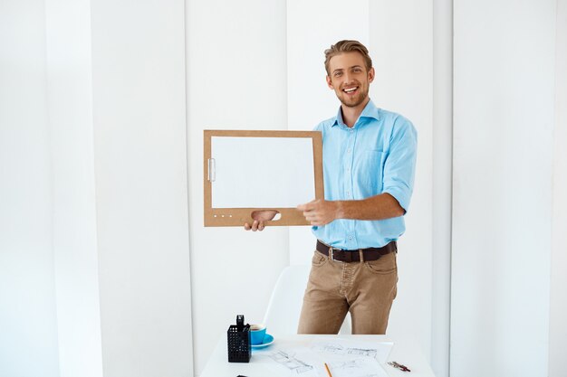 Young handsome cheerful smiling businessman standing at table holding wooden clipboard with white sheet. Light modern office interior 