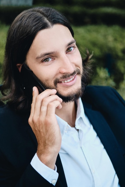 Young handsome confident bearded businessman joyfully looking in camera while talking on cellphone in restaurant outdoor