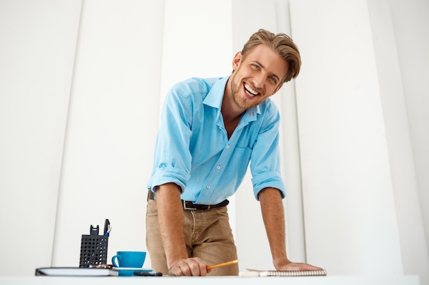 Young handsome confident cheerful businessman working standing at table over notepad looking smiling. White modern office interior.