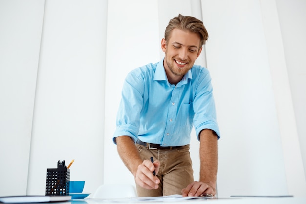 Бесплатное фото Молодой красивый уверенно жизнерадостный усмехаясь бизнесмен работая положение на эскизе чертежа таблицы. белый современный офисный интерьер