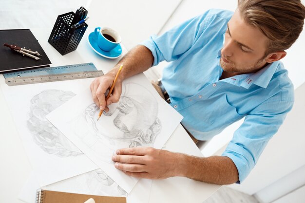 Young handsome confident pensive businessman sitting at table with pencil drawing portrait White modern office interior wall.