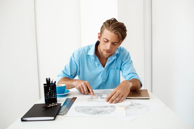 Young handsome confident pensive businessman sitting at table with pencil drawing portrait. White modern office interior.