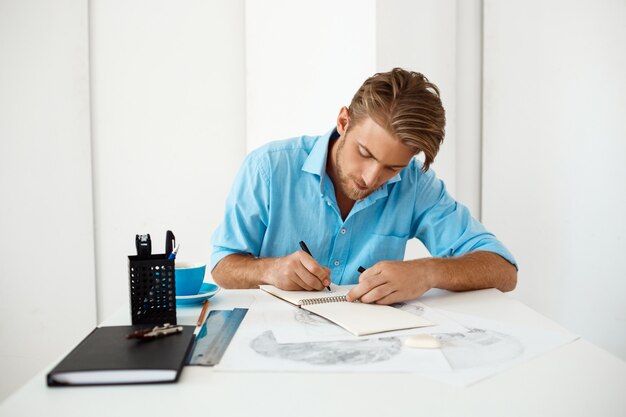Молодой красивый уверенно задумчивый бизнесмен сидя на таблице с портретом чертежа карандаша. Белый современный офисный интерьер.