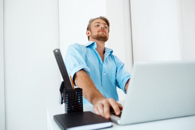 Young handsome confident pensive businessman sitting at table working on laptop with cup of coffee aside. White modern office interior 