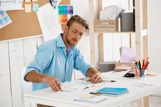 Young handsome confident pensive businessman working sitting at table looking through papers. White modern office interior 
