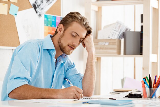 Young handsome confident pensive businessman working sitting at table writing in notepad. White modern office interior 