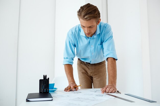 Young handsome confident pensive businessman working standing at table drawing sketch. White modern office interior 